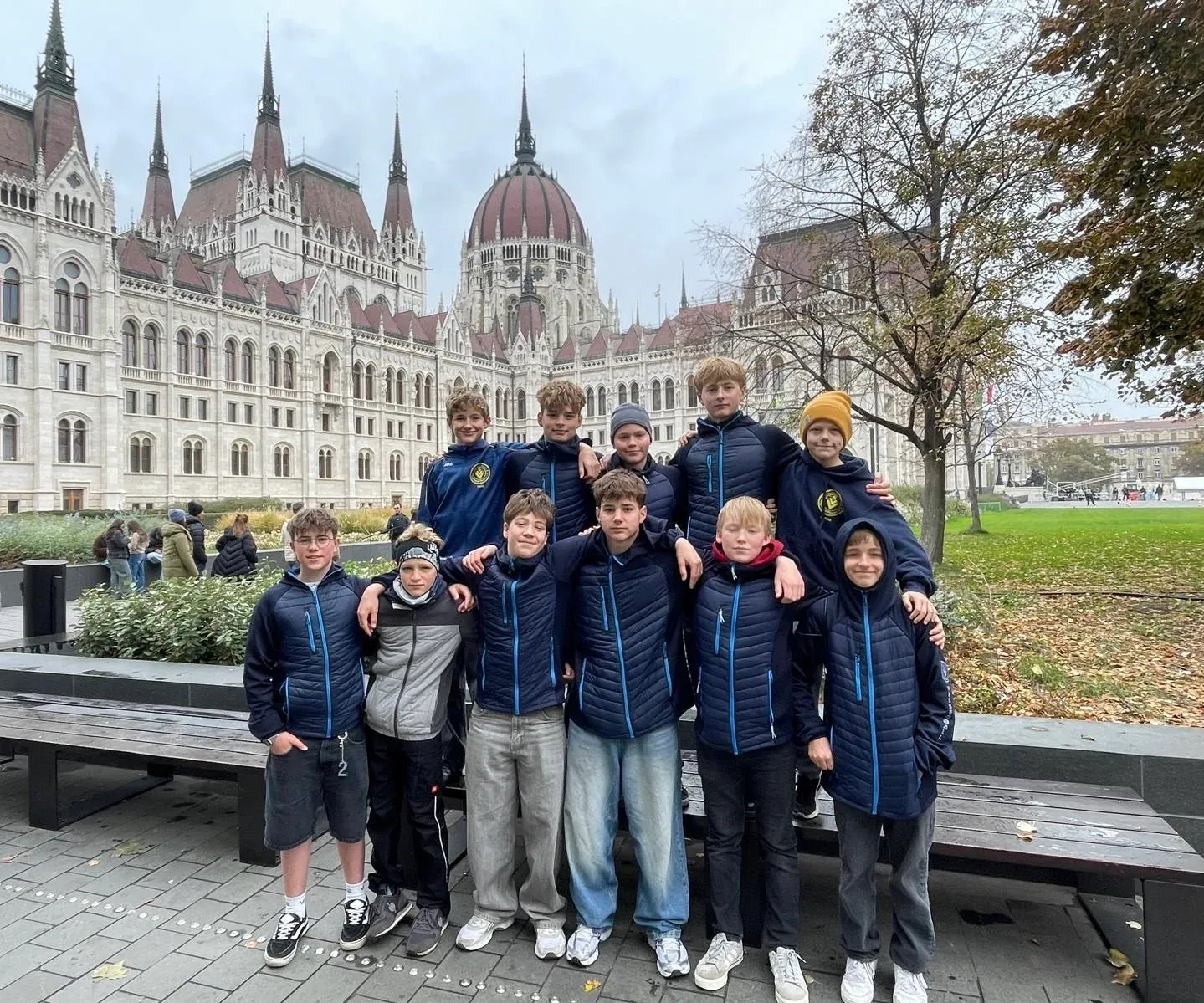 Elf Jugendliche in dunkelblauen Vereinsjacken posieren als Gruppe vor dem Parlamentsgebäude in Budapest.