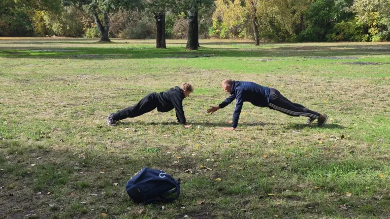 Ein Jugendlicher und ein Mann stützen sich auf einer Parkwiese im Liegestütz gegenüber, vorne liegt ein dunkelblauer Rucksack.