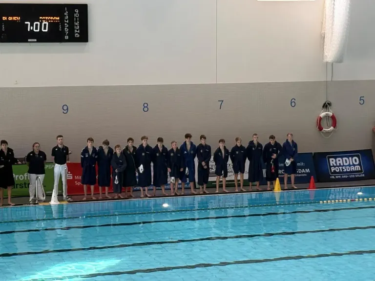 Wasserballmannschaft in dunkelblauen Bademänteln steht am Beckenrand, darüber Anzeigetafel „Plauen – Potsdam“ mit 7:00.