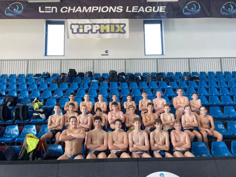 Jugendliche Wasserballer in Badehosen sitzen mit verschränkten Armen auf blauen Tribünensitzen unter Banner „LEN Champions League“.