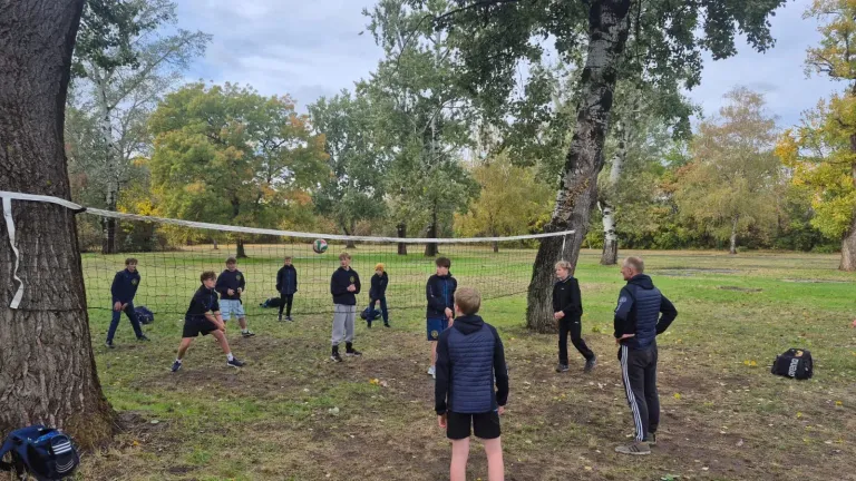 Jugendliche in dunkler Vereinskleidung spielen Volleyball an einem zwischen Bäumen gespannten Netz auf einer herbstlichen Wiese.